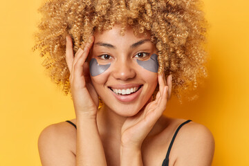 Wall Mural - Headshot of positive young woman with curly hair applies beauty patches under eyes to reduce puffiness smiles toothily has healthy smooth skin isolated over yellow background. Wellness concept