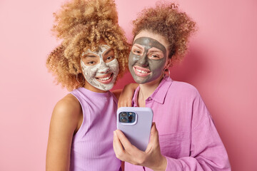 Wall Mural - Positive young female friends with curly hair applies beauty mask for skin treatment use mobile phone dressed in casual clothes take selfie isolated over pink background. Facial care concept