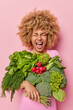 © wayhome.studio  - Indoor shot of curly haired young woman carries fresh green vegetables being vegetarian eats healthy organic food keeps to diet exclaims louldy isolated over pink background. Seasonal vitamins