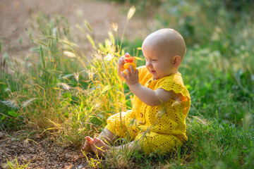 little cute happy baby girl sitting on the green grass on a sunny summer day dressed in knitted cotton yellow jumpsuit.