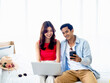 © tete_escape - Happy Asian couple, young man and woman in casual looking at smart mobile phone screen while using and working with laptop computer together, video call with their friends in bright room.