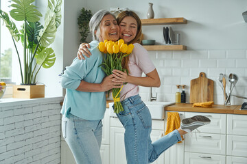 Wall Mural - Happy senior mother receiving a bunch of tulips from her daughter while standing at the kitchen