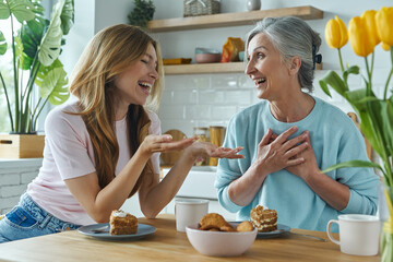 Wall Mural - Happy mother and her adult daughter communicating while enjoying sweet food at the kitchen