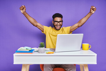 excited man looking at laptop and gesturing while sitting at the desk against purple background