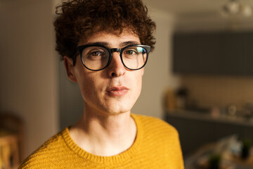 Closeup portrait of handsome young man with curly hair, brown eyes and in glasses looking confidently at camera standing at home