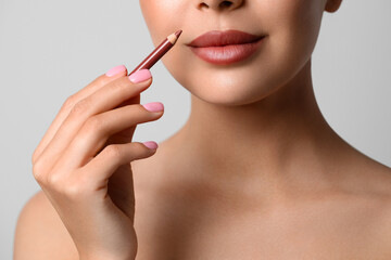 Poster - Young woman applying beautiful nude lip pencil on light grey background, closeup