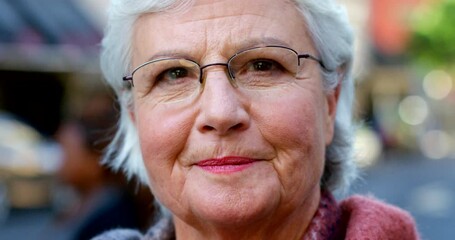 Poster - Portrait of a senior woman wearing glasses and smiling while out in the urban city. Face of a wrinkled elderly lady with grey hair standing outside. Pensioner traveling and enjoying her retirement