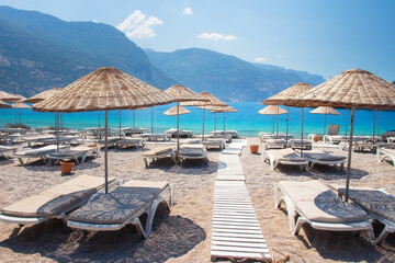 Fototapete - Landscape of beautiful coastline in Oludeniz with straw umbrellas. View from beach on blue sea and mountains, amazing rest in Turkey. Beginning of day on beach without people.