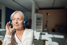 Smiling Senior Woman With Eyes Closed Listening Music Through Wireless Headphones In Office