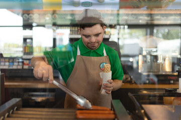 Wall Mural - Happy waiter with Down syndrome standing by counter and prepairing hot-dog to a customer in cafe at gas station.