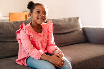 Wall Mural - African american little girl smiling while sitting on sofa at home