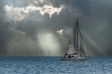 Dozens Of Off-shore, Industrial Wind Turbines Generating Environmentally Friendly Electricity With A Sailboat In The Foreground. 