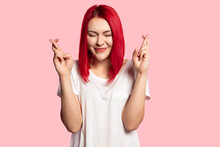 Portrait Of Lovely Girl Posing With Crossed Fingers And Waiting For Her Wish To Come True. Adorable Model With Bright Red Hairstyle. Happiness And Lifestyle Concept. Isolated On Pink