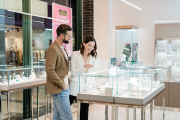 Smiling young woman pointing at jewelry near bearded boyfriend in store.