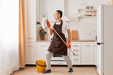 Wall Mural - Full length photo of young guy cleaning kitchen, having fun, listening music and singing song, using mop as microphone, dancing, being in good mood while cleaning his apartment.