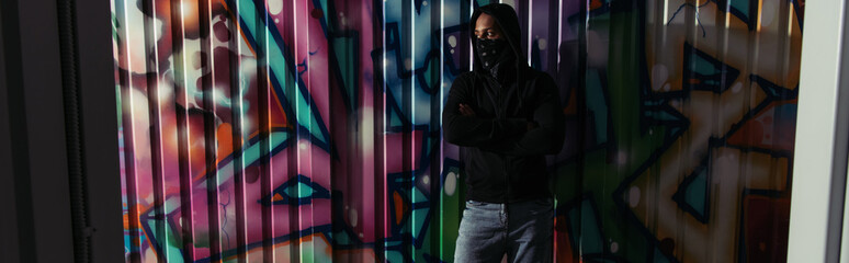 Wall Mural - African american hooligan in hoodie and face mask standing near graffiti on wall, banner