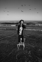 Romantic Girl In Stylish Formal Suit, Shirt And Hat Stands On White Chair In The Water Of Sea Or Ocean. Creative Romantic Moody Black And White Photo Loneliness Puberty Metaphor. Seagulls Fly In Sky