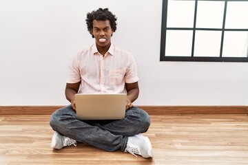 Canvas Print - African man with curly hair using laptop sitting on the floor sticking tongue out happy with funny expression. emotion concept.