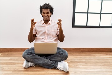 Poster - African man with curly hair using laptop sitting on the floor gesturing finger crossed smiling with hope and eyes closed. luck and superstitious concept.