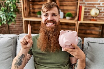 Poster - Redhead man holding piggy bank smiling with an idea or question pointing finger with happy face, number one