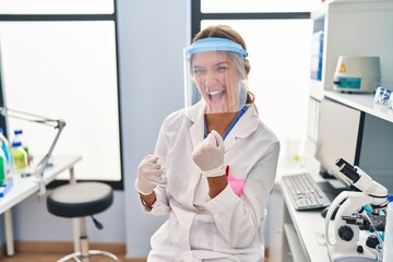 Canvas Print - Young blonde woman working at scientist laboratory wearing face mask celebrating surprised and amazed for success with arms raised and eyes closed