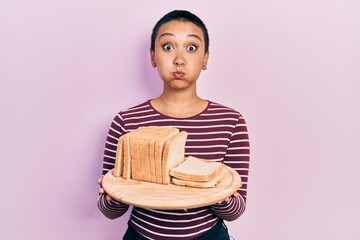 Canvas Print - Beautiful hispanic woman with short hair holding sandwich bread puffing cheeks with funny face. mouth inflated with air, catching air.