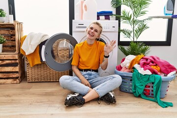 Poster - Young blonde woman doing laundry sitting by washing machine waiving saying hello happy and smiling, friendly welcome gesture