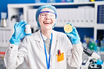 Canvas Print - Beautiful woman working at scientist laboratory holding pills and lemon smiling and laughing hard out loud because funny crazy joke.