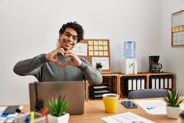 Canvas Print - Young hispanic man wearing business style sitting on desk at office smiling in love doing heart symbol shape with hands. romantic concept.