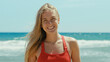 Portrait of beautiful young woman in red swimsuit stand on empty sunny beach, look into camera with confidence. Happy authentic smile. Summertime concept. Warm breeze in hair