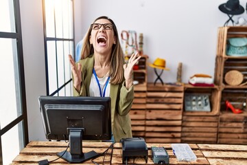 Young hispanic woman working as manager at retail boutique crazy and mad shouting and yelling with aggressive expression and arms raised. frustration concept.