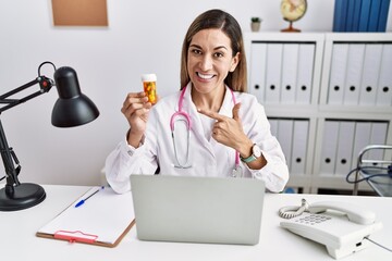 Canvas Print - Young hispanic woman wearing doctor uniform holding pills at the clinic cheerful with a smile on face pointing with hand and finger up to the side with happy and natural expression