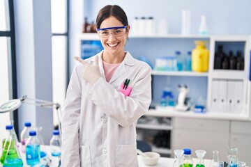 Sticker - Young brunette woman working at scientist laboratory smiling cheerful pointing with hand and finger up to the side