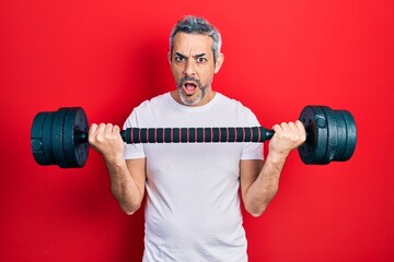 Poster - Handsome middle age man with grey hair wearing sportswear using dumbbells in shock face, looking skeptical and sarcastic, surprised with open mouth