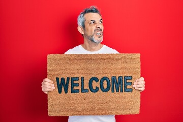 Sticker - Handsome middle age man with grey hair holding welcome doormat angry and mad screaming frustrated and furious, shouting with anger. rage and aggressive concept.