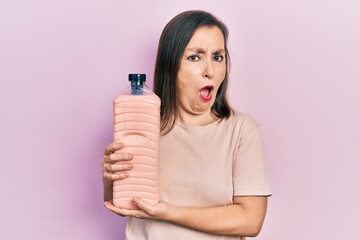 Poster - Middle age hispanic woman holding detergent bottle in shock face, looking skeptical and sarcastic, surprised with open mouth