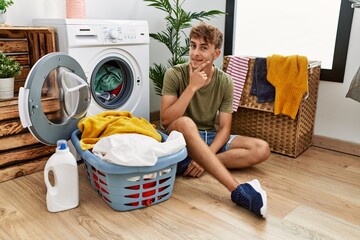 Sticker - Young caucasian man putting dirty laundry into washing machine looking confident at the camera smiling with crossed arms and hand raised on chin. thinking positive.