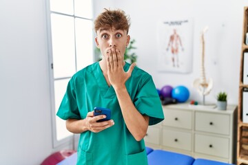 Sticker - Young caucasian physio man using smartphone at medical clinic covering mouth with hand, shocked and afraid for mistake. surprised expression