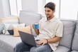 © Krakenimages.com - Young hispanic man using laptop and drinking coffee sitting on sofa at home
