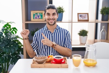 Wall Mural - Hispanic man with long hair sitting on the table having breakfast smiling and looking at the camera pointing with two hands and fingers to the side.