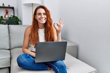 Poster - Young redhead woman using laptop at home doing ok sign with fingers, smiling friendly gesturing excellent symbol