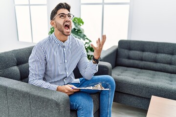 Sticker - Young psychologist man at consultation office crazy and mad shouting and yelling with aggressive expression and arms raised. frustration concept.