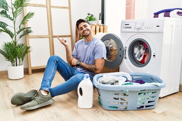 Wall Mural - Young hispanic man putting dirty laundry into washing machine with a big smile on face, pointing with hand finger to the side looking at the camera.