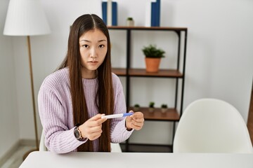 Wall Mural - Young chinese girl holding pregnancy test sitting on the table at home.
