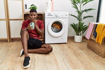 Canvas Print - Young african american man using smartphone waiting for washing machine laughing and embarrassed giggle covering mouth with hands, gossip and scandal concept