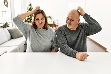 Sticker - Middle age hispanic couple sitting on the table by christmas tree confuse and wonder about question. uncertain with doubt, thinking with hand on head. pensive concept.