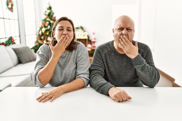 Wall Mural - Middle age hispanic couple sitting on the table by christmas tree bored yawning tired covering mouth with hand. restless and sleepiness.