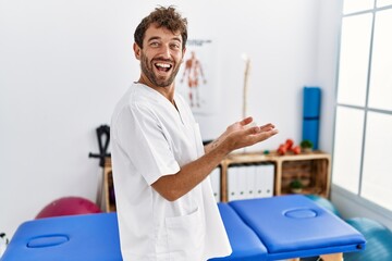 Poster - Young handsome physiotherapist man working at pain recovery clinic pointing aside with hands open palms showing copy space, presenting advertisement smiling excited happy