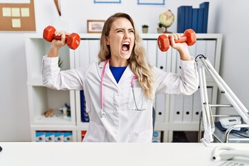 Sticker - Young beautiful doctor woman holding dumbbells for sport therapy angry and mad screaming frustrated and furious, shouting with anger. rage and aggressive concept.