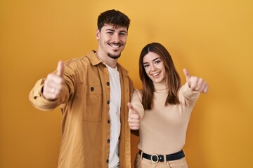 Young hispanic couple standing over yellow background approving doing positive gesture with hand, thumbs up smiling and happy for success. winner gesture.